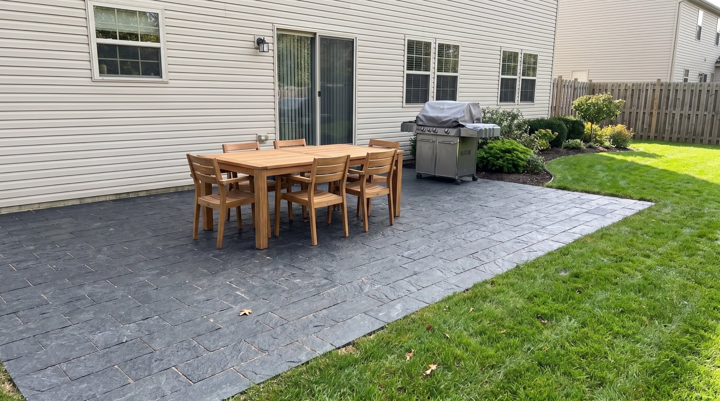 Dark splitface granite paver patio behind a house with a wooden dining table and a grill